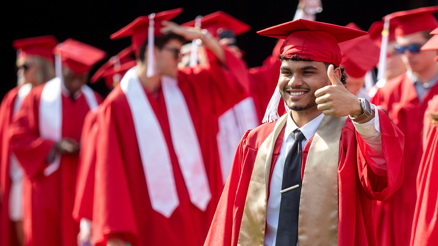 A graduating student, surrounded by other students in red caps and gowns, gives a thumbs up to someone in the crowd.