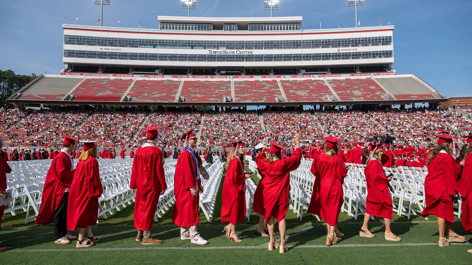 A line of students wearing red graduation caps and gowns stands on the field beside rows of seats at Carter-Finley Stadium.