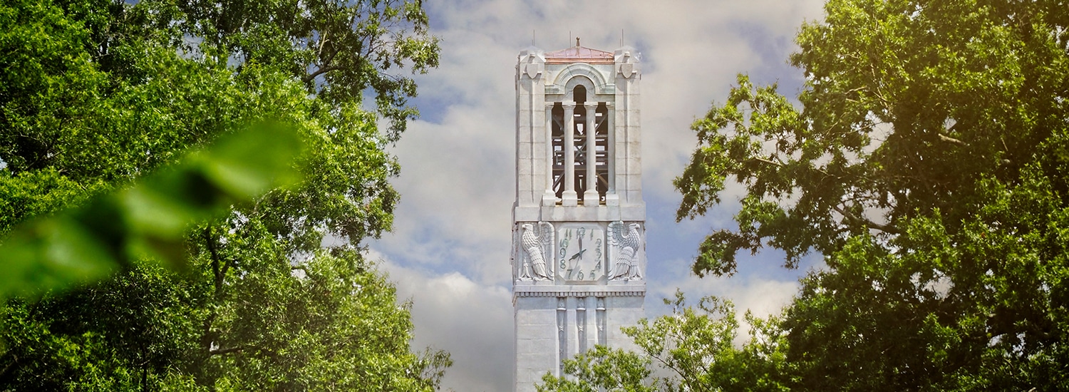The top of NC State's Memorial Belltower peeking through leafy trees.
