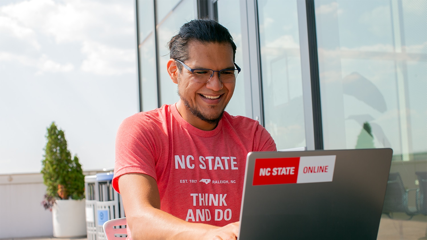 A man in an NC State T-shirt works at a laptop with a sticker reading "NC State Online."